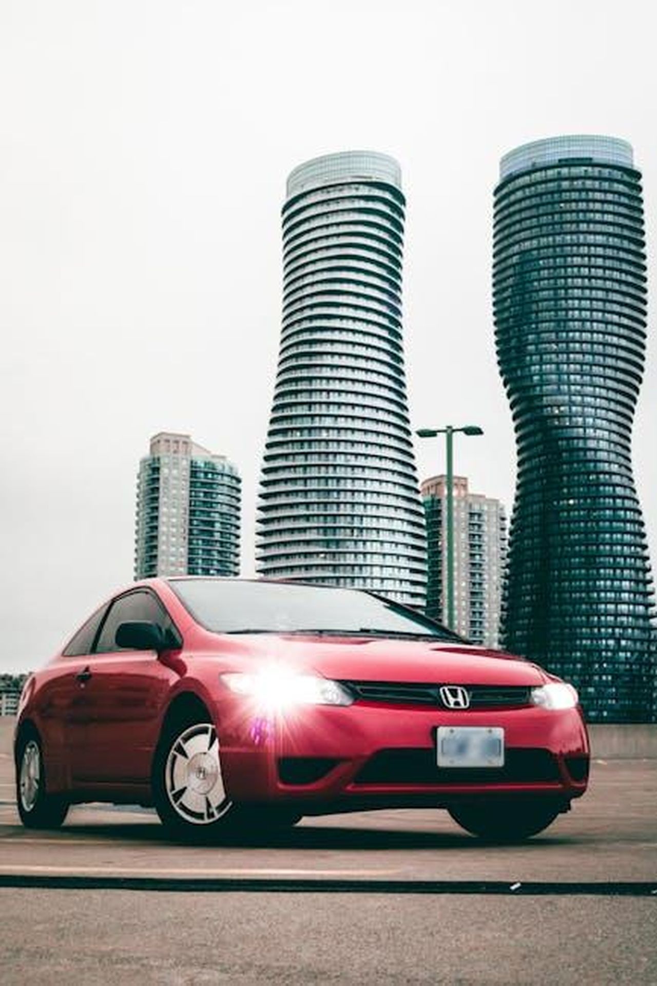 2025_Honda_Civic_Hybrid_efficienza_e_prestazioni_r4QRkiq1ng - Veicoli24 Source: Javon Swaby - pexels - Red Honda car parked in front of distinctive twisted towers in Mississauga, Canada.