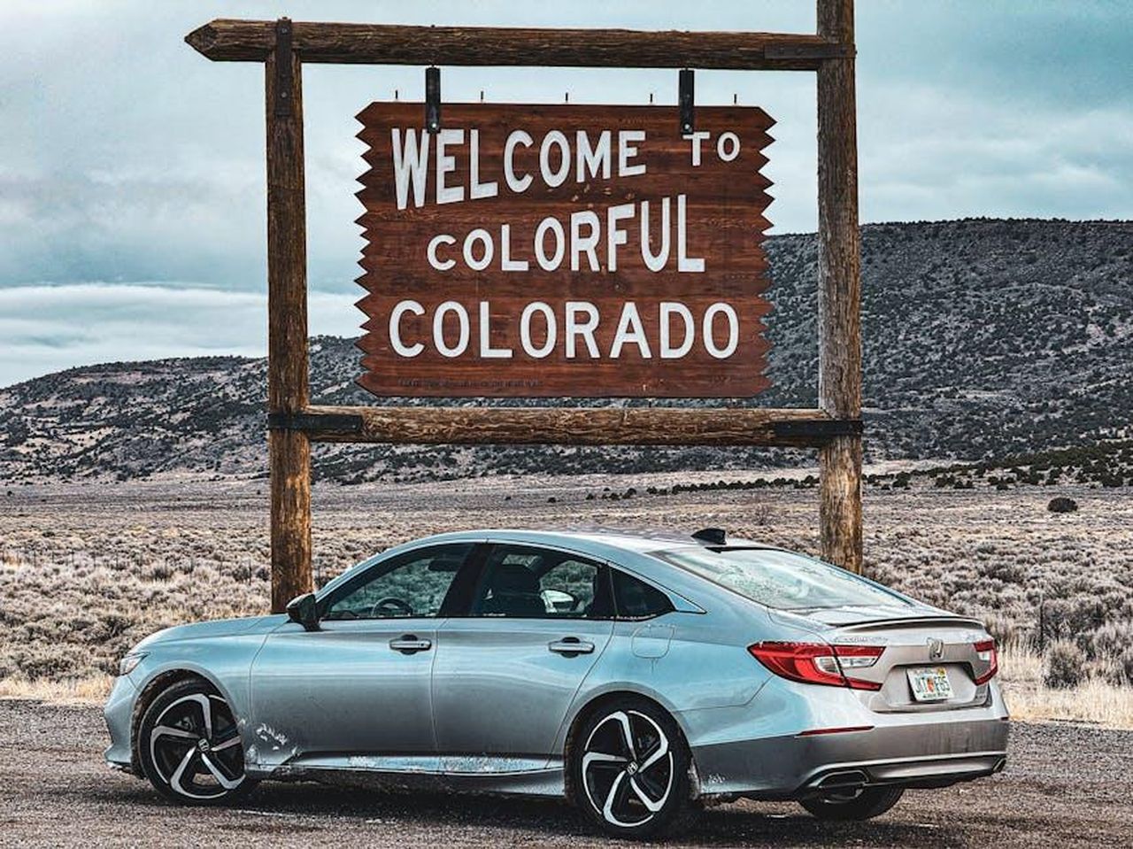Caratteristiche_e_specifiche_X170mef1Pi - Veicoli24 Source: HRK Gallery - pexels - A silver sedan parked by a scenic Welcome to Colorful Colorado sign on a cloudy day.