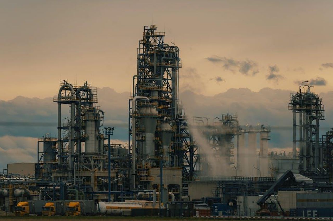 Carburante_ed_economia_h3XvJ6ELzG - Veicoli24 Source: Michael Pointner - pexels - A detailed view of a refinery's towers against a cloudy dusk sky, showcasing industrial architecture.