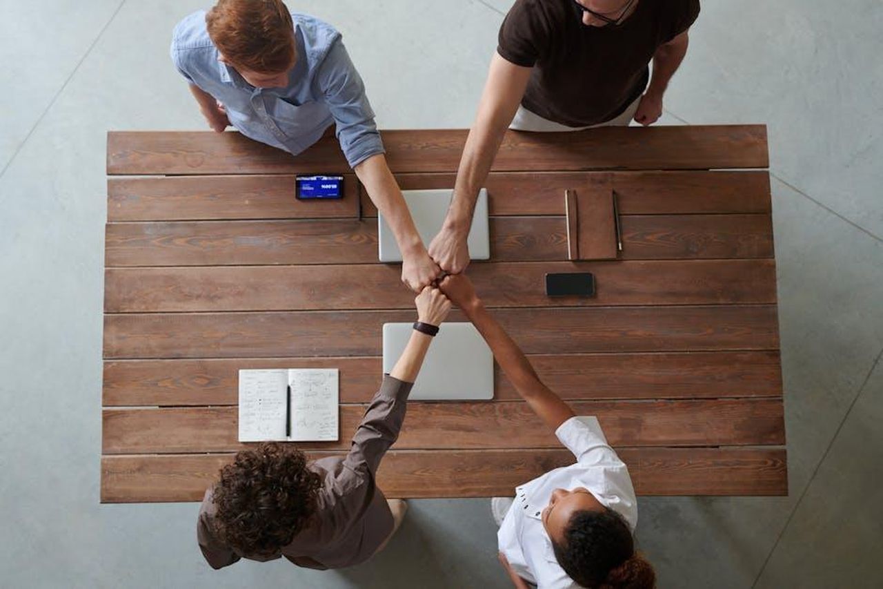 Commenti_dei_dirigenti_ak0zdAcpfb - Veicoli24 Source: fauxels - pexels - A diverse group of professionals engaging in a teamwork celebration with a fist bump over a wooden table indoors.
