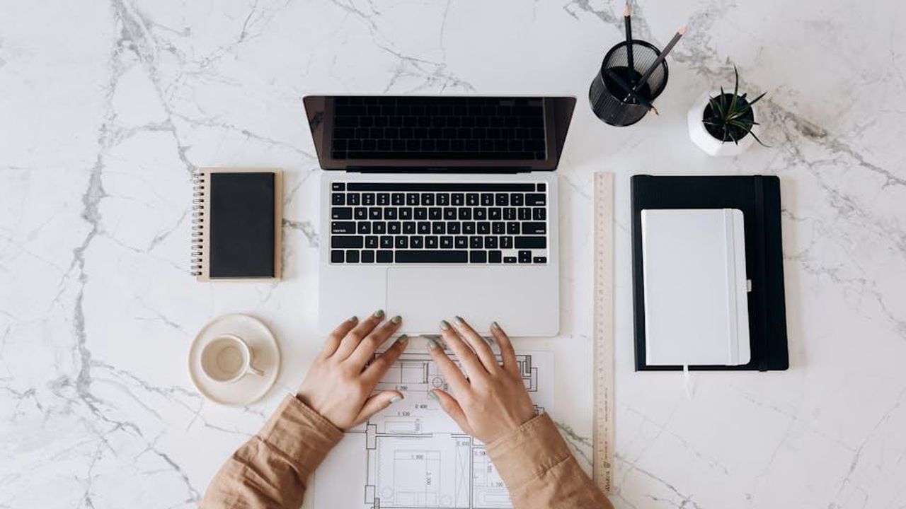 Commento_del_CEO_Graeme_Nieman_iWf3mIJHqv - Veicoli24 Source: Mikhail Nilov - pexels - Top view of a stylish home office desk with a laptop, planner, and coffee cup, showing hands on a blueprint.