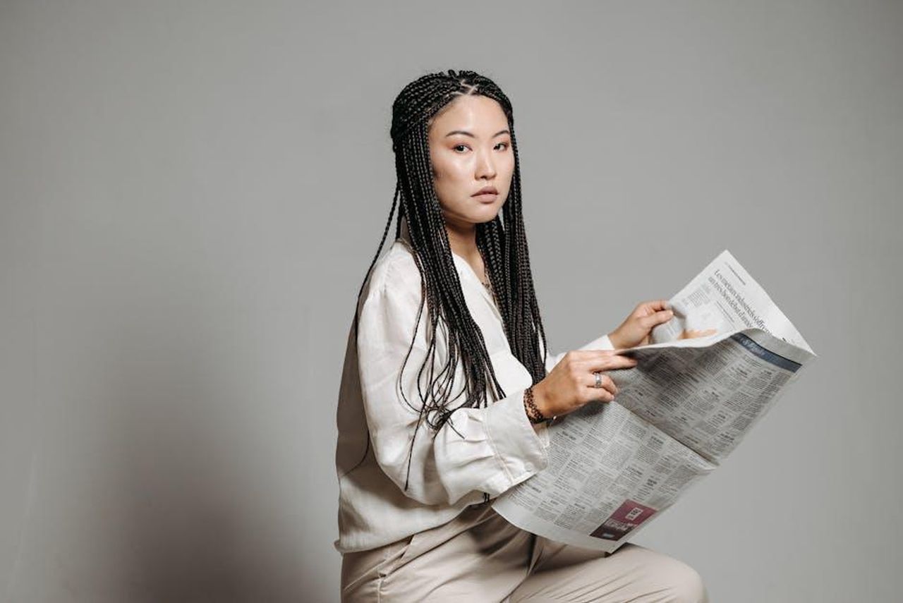 Contatti_Stampa_TCqYvP4QRJ - Veicoli24 Source: Pavel Danilyuk - pexels - Asian woman with braided hair reads a newspaper, wearing a button-down shirt in a studio setting.
