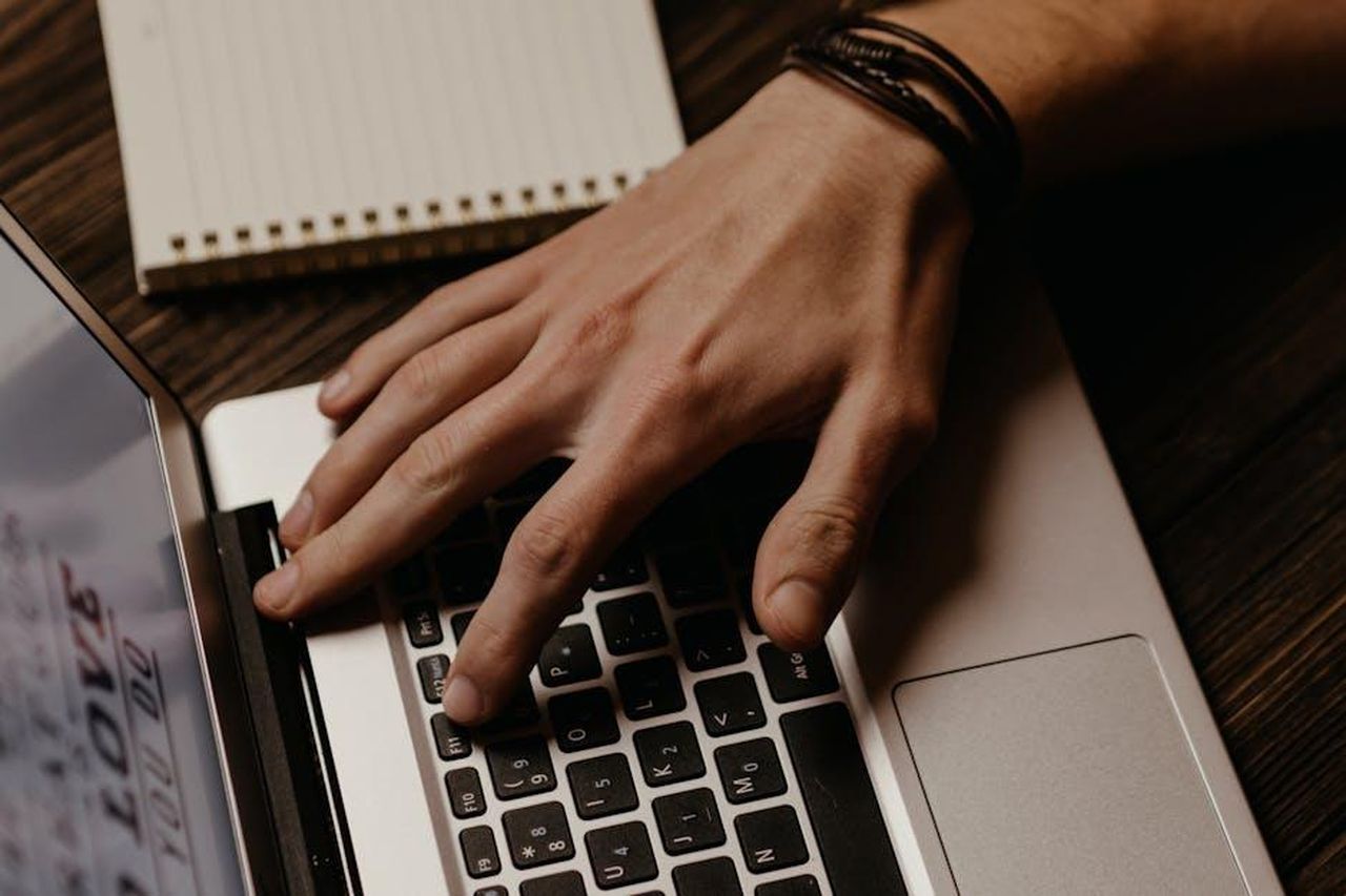 Contatti_stampa_e_autore_m5JQ7VilNz - Veicoli24 Source: Polina ⠀ - pexels - A close-up image showing a hand typing on a laptop with a notebook in the background.