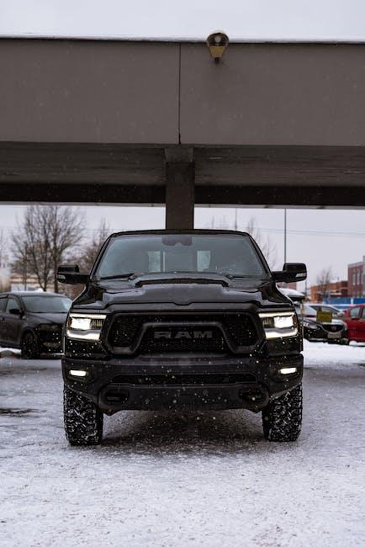 Cosa_ci_piace_della_Ram_1500_v5ZidnBzAL - Veicoli24 Source: Vitali Adutskevich - pexels - Front view of a black Dodge Ram 1500 Rebel parked under a bridge in a snowy urban area.