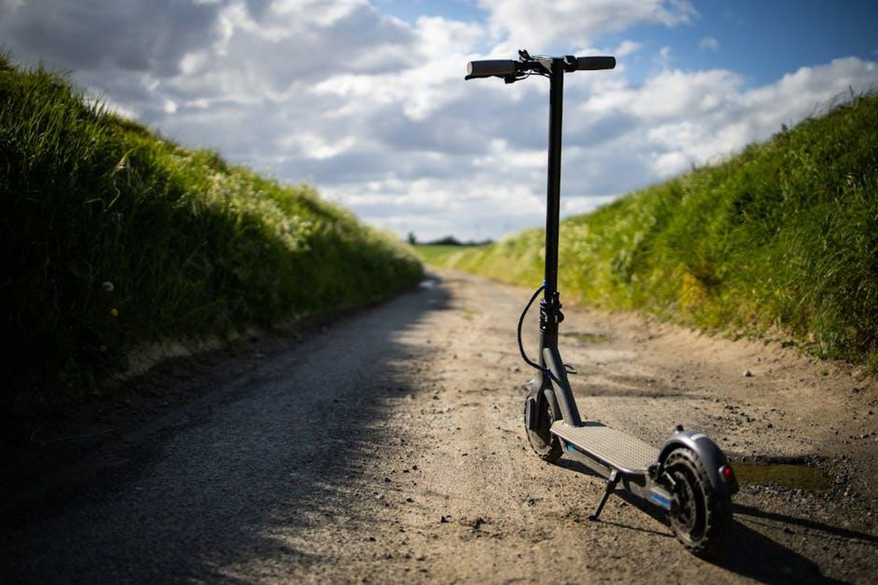 Crescita_dei_veicoli_elettrici_e_ibridi_rPQF75ltgm - Veicoli24 Source: Fabien Burgue - pexels - A solitary electric scooter on a deserted rural pathway with lush green grass and a bright blue sky, evoking freedom.