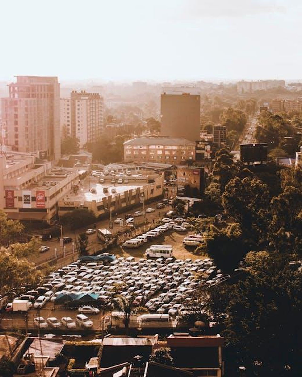 Espansione_delle_operazioni_di_GMG_2wCaCKtl5u - Veicoli24 Source: Mwabonje Ringa - pexels - Aerial view of a bustling cityscape featuring buildings, parked vehicles, and trees in warm daylight.