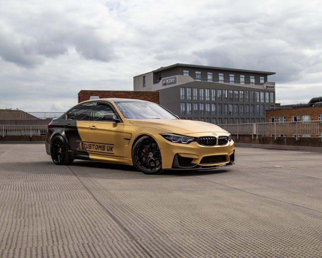 Nuova_divisione_di_Gordon_Murray_Special_Vehicles_9GOCX8bcPR - Veicoli24 Source: d30visuals . - pexels - A sleek gold car parked in an open lot with modern buildings in the background.