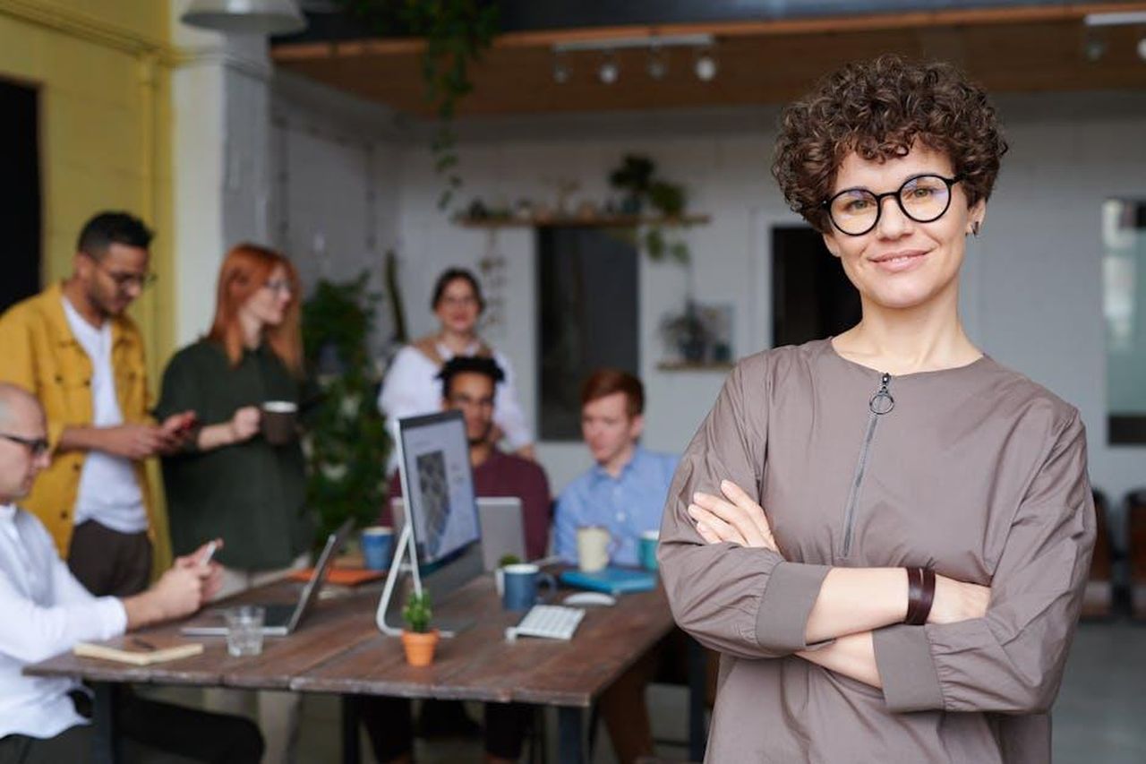 Nuovo_CEO_e_rilancio_previsto_iwSnhXGQZx - Veicoli24 Source: fauxels - pexels - Smiling businesswoman with curly hair stands confidently in a modern office space with colleagues.
