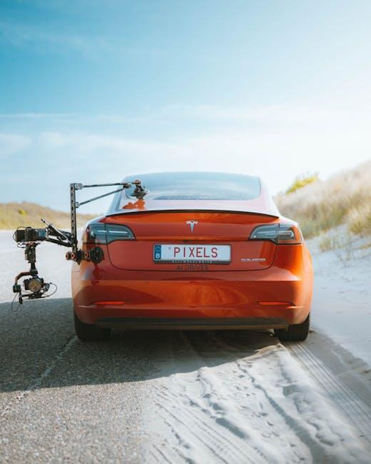 Piani_futuri_di_Toyota_per_la_produzione_di_veicoli_elettrici_W41z6NxkHF - Veicoli24 Source: Jérémy Glineur - pexels - Red Tesla Model 3 with a camera rig, driving on a sandy road under a clear sky.