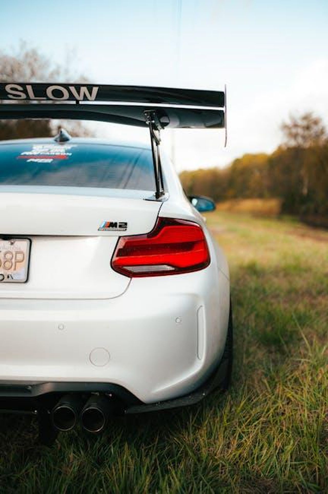 Pilota_ufficiale_BMW_M_dal_2023_zk4jhp7nq5 - Veicoli24 Source: Luke Miller - pexels - Rear view of a white sportscar with a large spoiler, parked outdoors on grass.