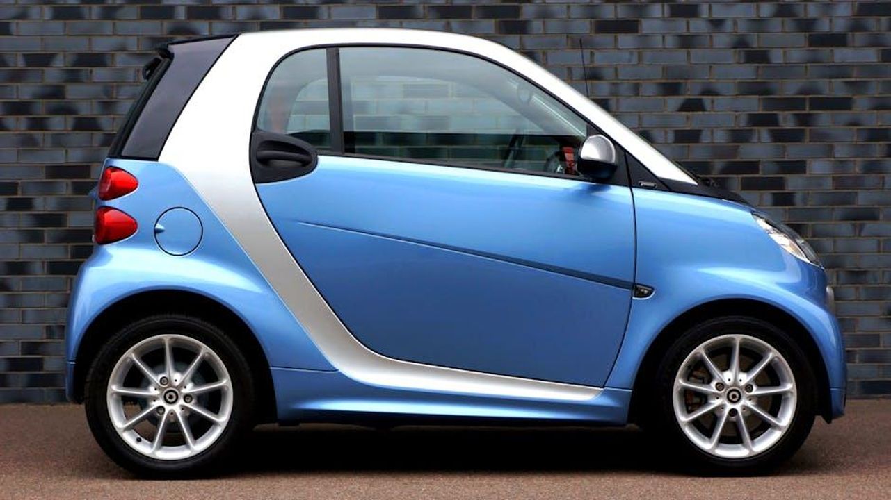 Progetti_simili_nel_settore_automobilistico_9i8dIJjQtN - Veicoli24 Source: Mike Bird - pexels - Side view of a modern blue smart car against a brick wall, showcasing compact design.