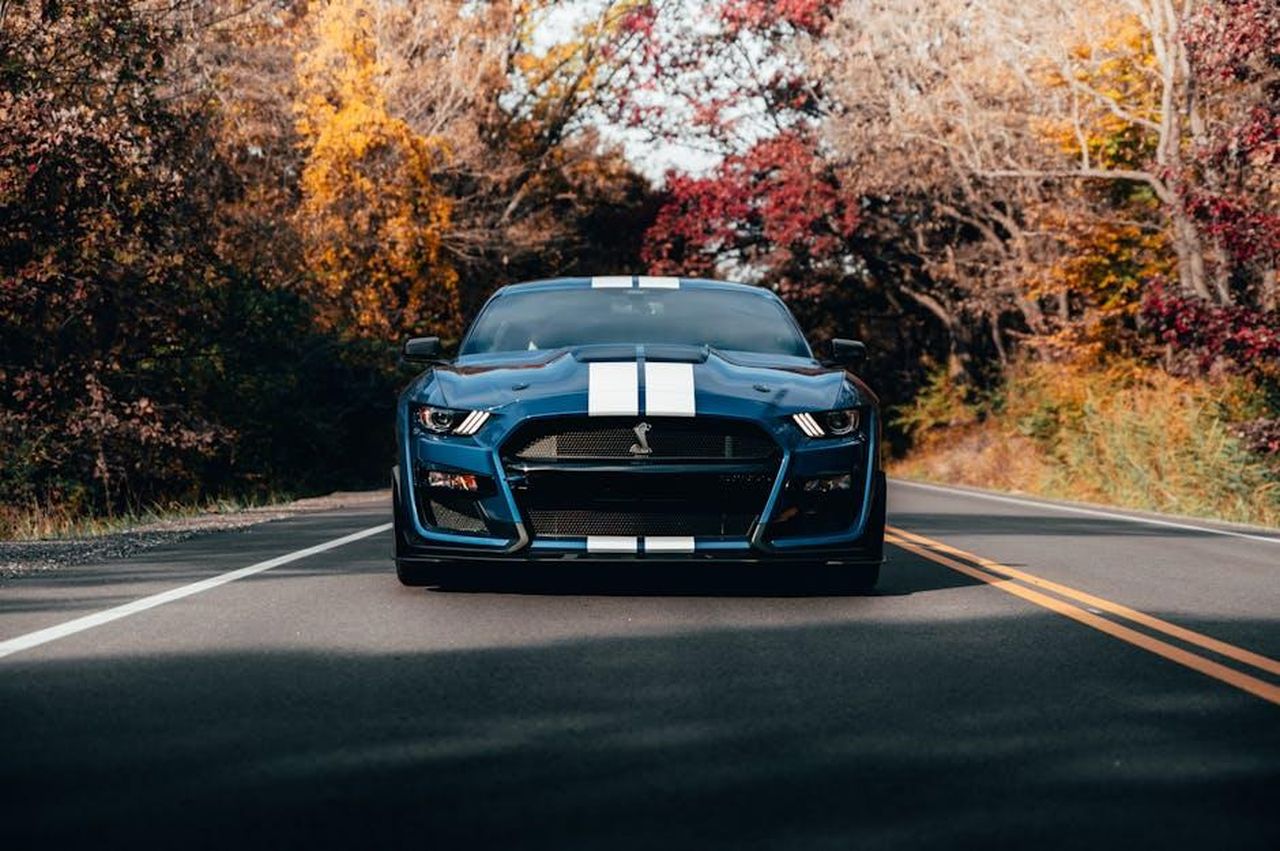 Richiamo_del_Ford_Transit_Trail_nel_2024_UDcBKXo0i5 - Veicoli24 Source: Max Andrey - pexels - Front view of a blue Ford Mustang driving on a road surrounded by autumn trees with colorful foliage.
