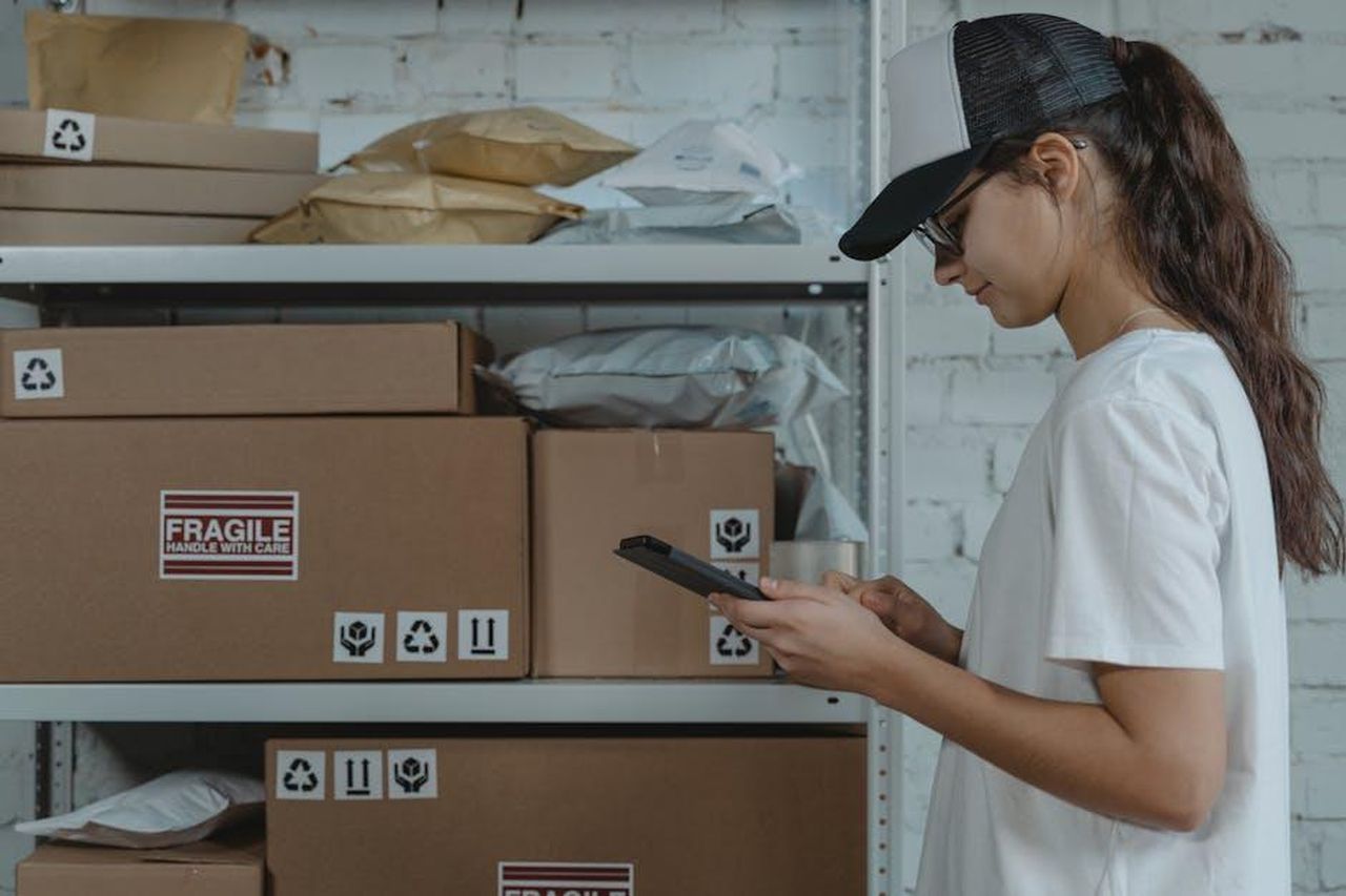 Valore_delle_esportazioni_nel_2024_e_importanza_del_commercio_libero_xHMx2MmI2i - Veicoli24 Source: Tima Miroshnichenko - pexels - A female courier in uniform managing packages with a device in a storage room.