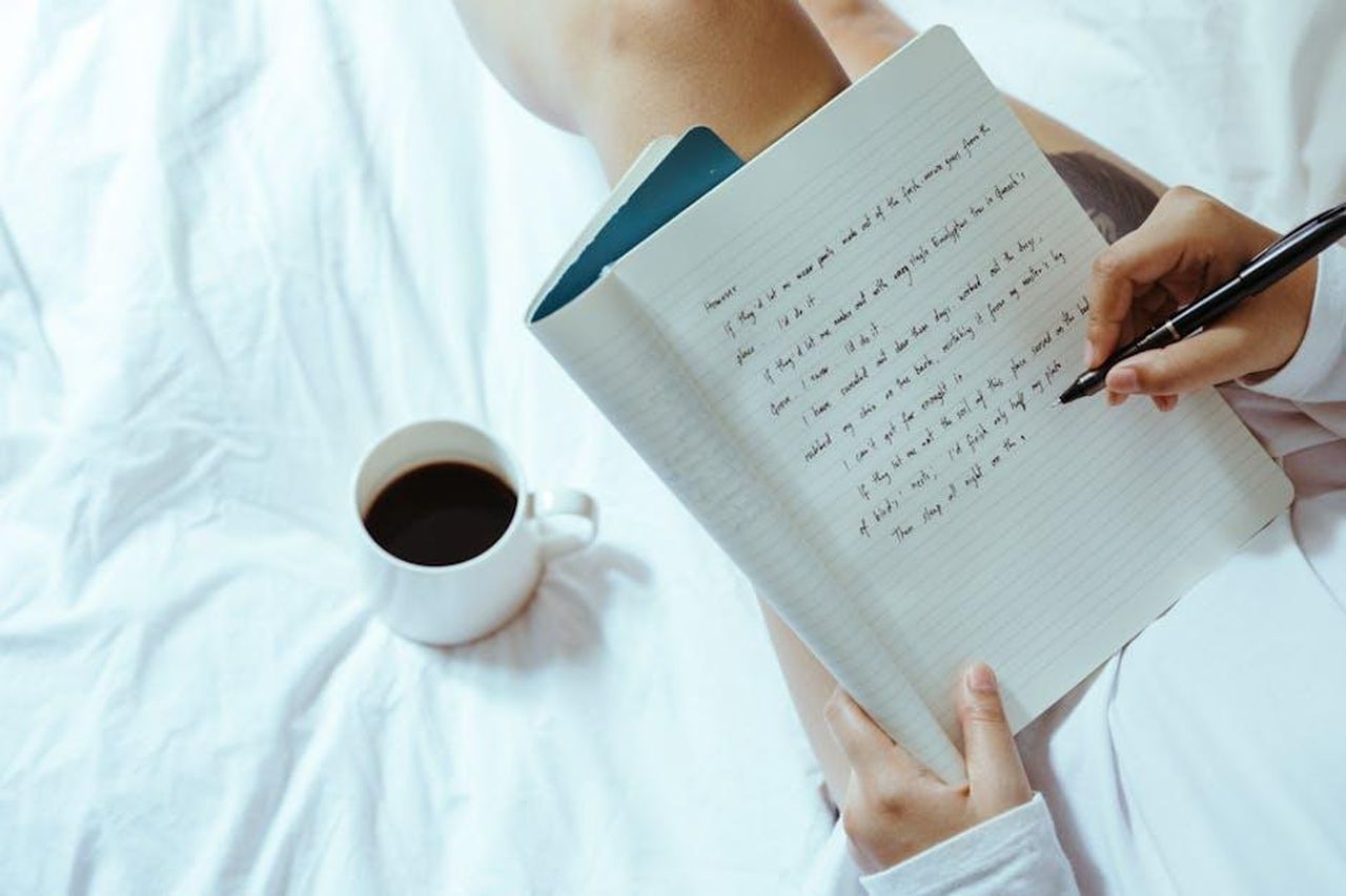 Autore_laARmGRKVZ - Veicoli24 Source: Ketut Subiyanto - pexels - Top view of unrecognizable woman sitting on bed with legs near cup of coffee and writing on notepad with pen while resting at home