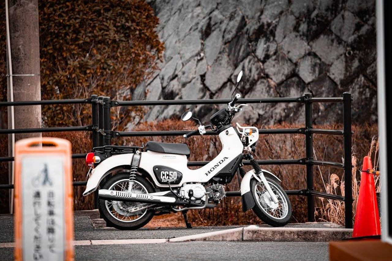 Confronto_tra_Honda_Prologue_2025_e_2024_RQS3wdEZWn - Veicoli24 Source: Dinesh Silwal - pexels - A detailed photograph of a classic white Honda motorcycle parked by a fence in an outdoor setting.