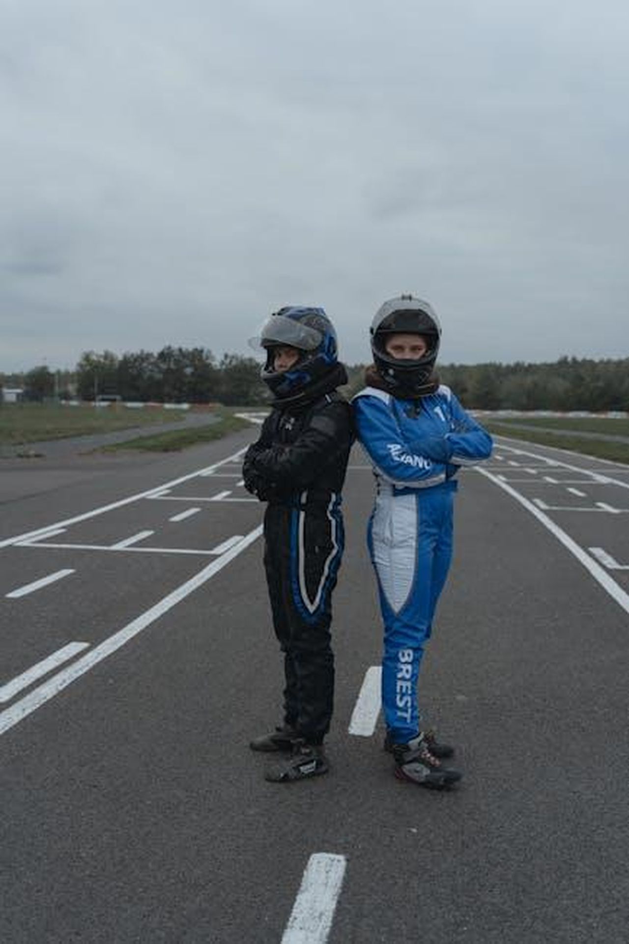Dichiarazioni_dei_piloti_Vasser_Sullivan_AOtinKGFTW - Veicoli24 Source: Tima Miroshnichenko - pexels - Two race drivers in helmets and suits posing back to back on a track, showcasing motorsport readiness.