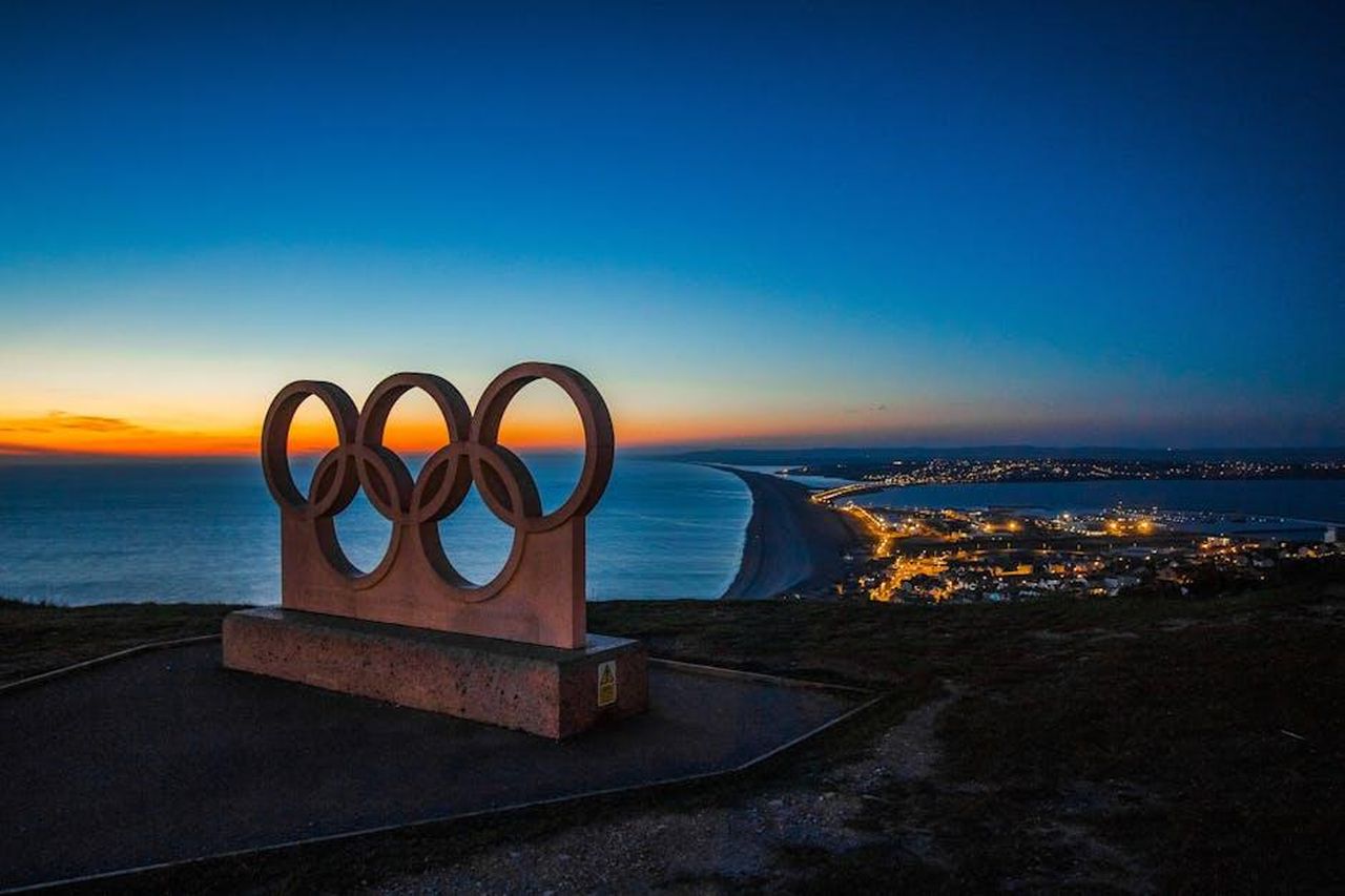 Impegno_di_Stellantis_per_i_Giochi_Olimpici_e_Paralimpici_PkUSTH4GHl - Veicoli24 Source: Pixabay - pexels - Scenic sunset view of the Olympic rings monument by the sea with city lights in the distance.