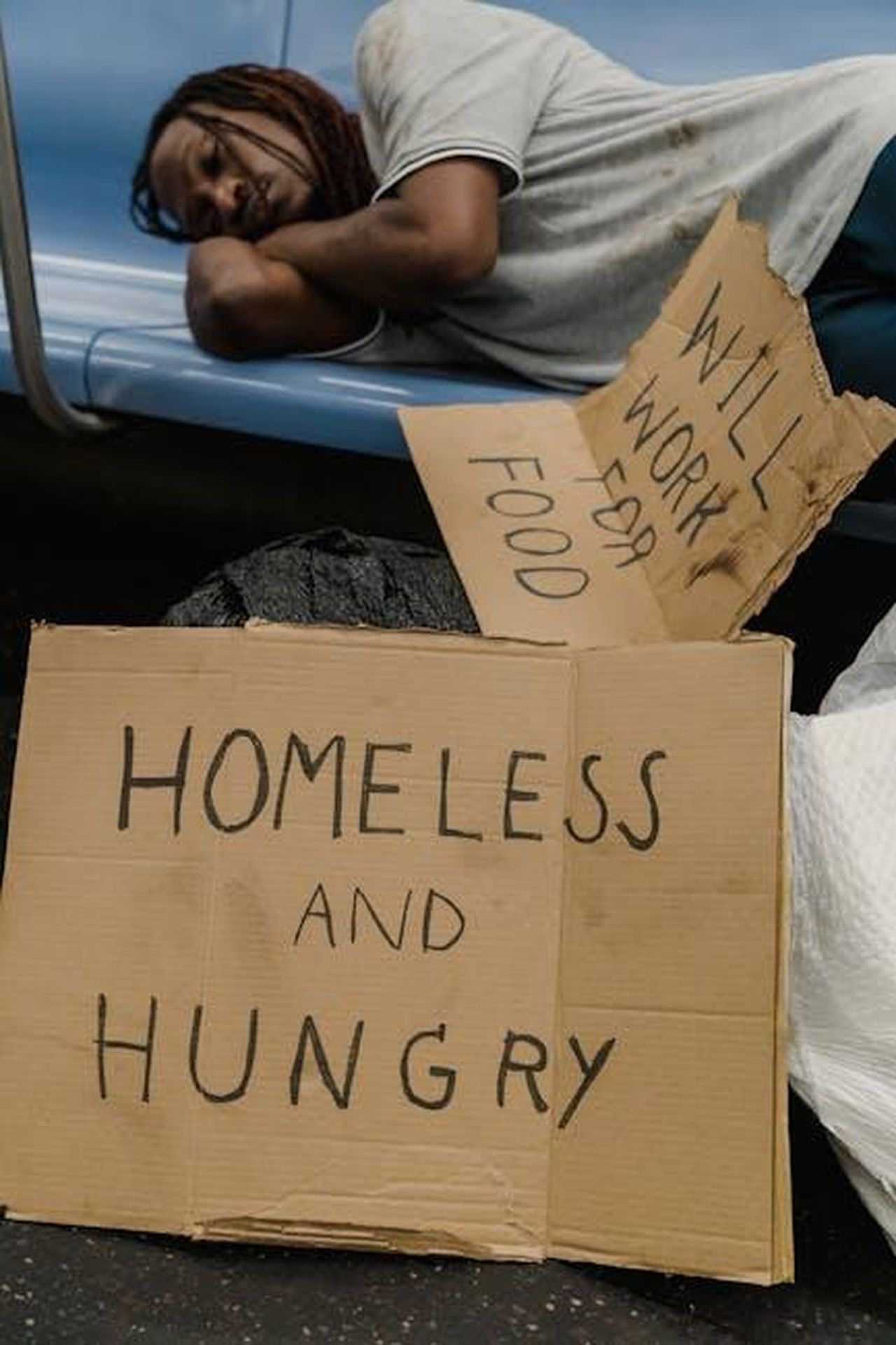 Importanza_delle_stazioni_di_ricarica_per_la_comunit_u7jOnokSD8 - Veicoli24 Source: Timur Weber - pexels - Man sleeping on subway bench with cardboard signs highlighting homelessness.