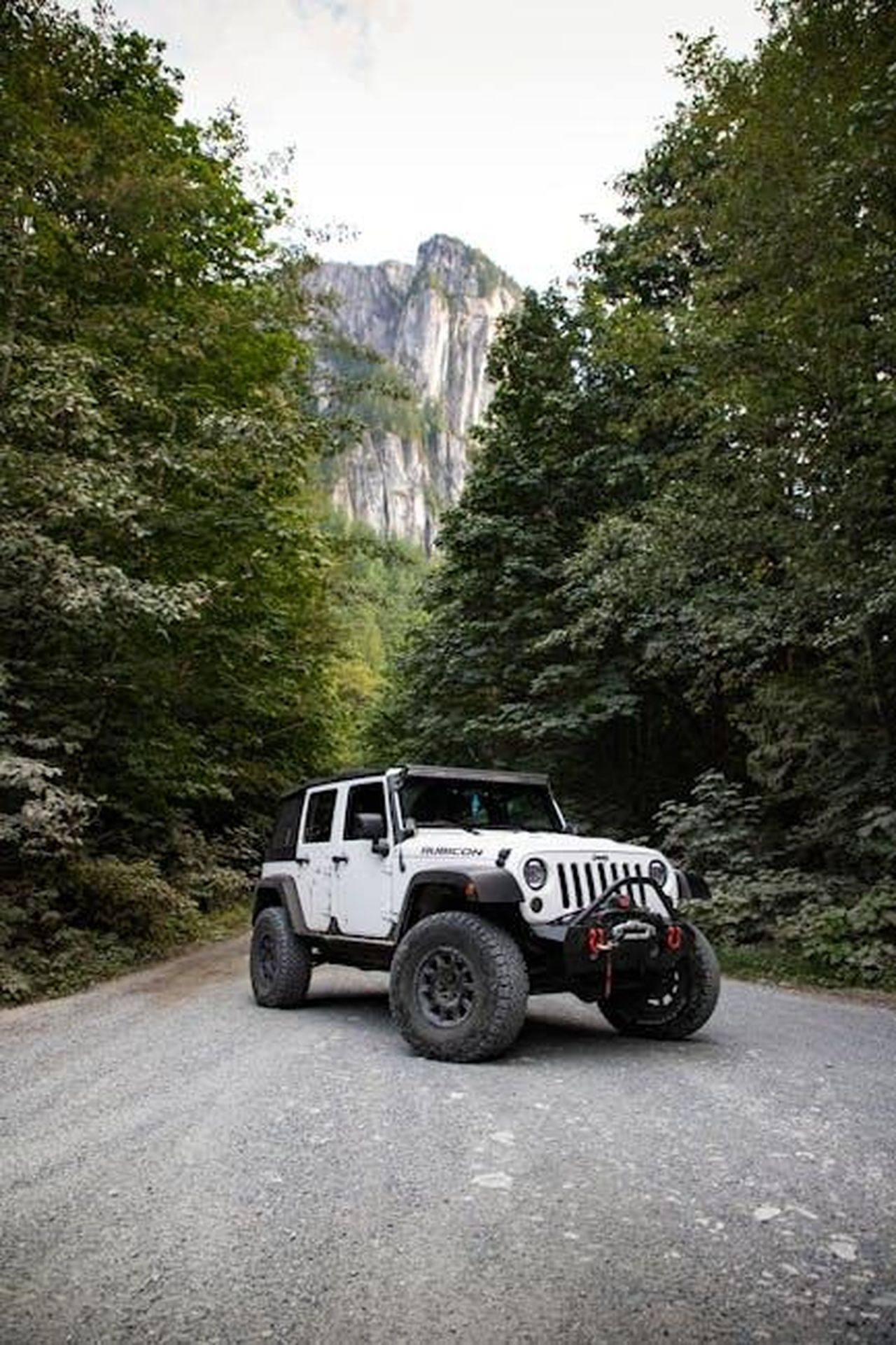 Jeep_Wrangler_4xe_licona_elettrificata_del_brand_bkrmkwKBwP - Veicoli24 Source: Zsolt Joo - pexels - A rugged Jeep Wrangler on a gravel road surrounded by lush forest and towering mountains.