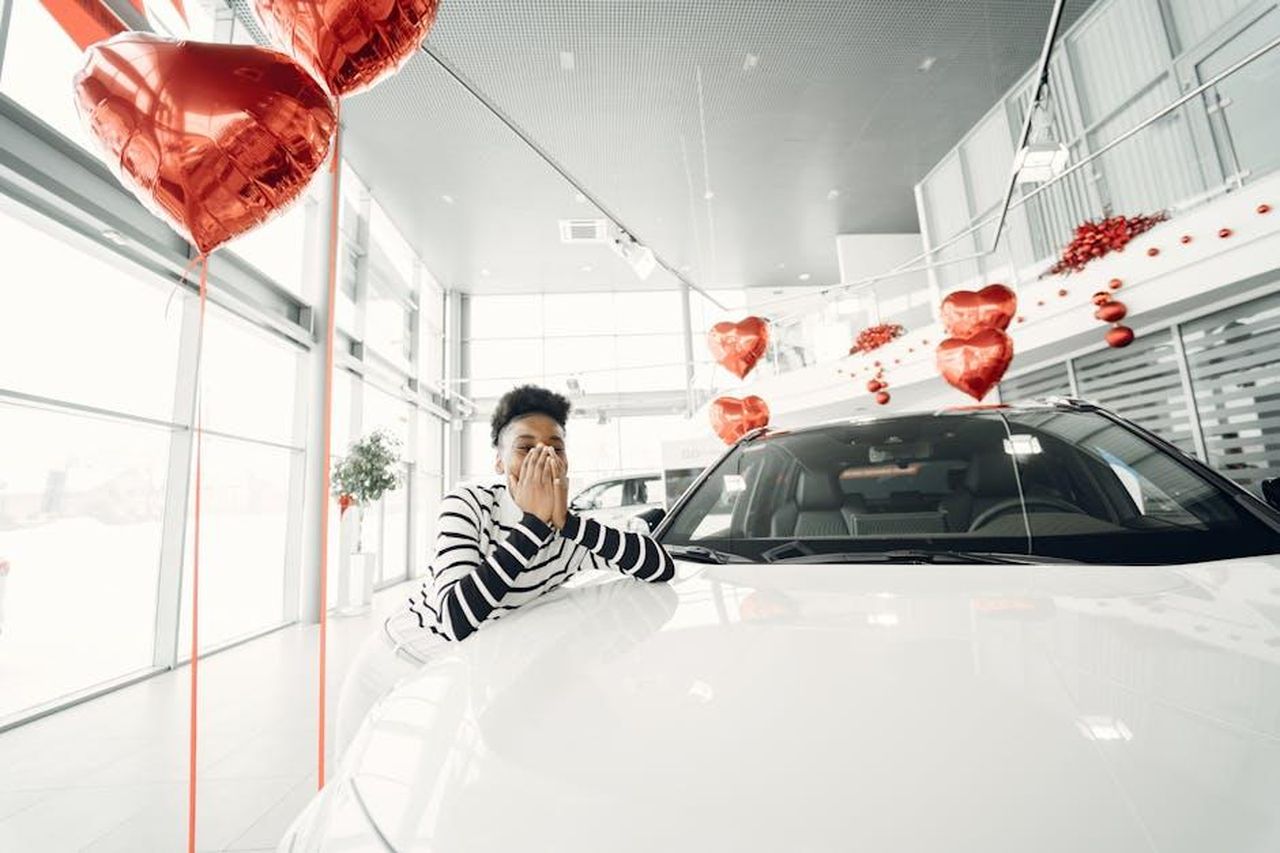 Opzioni_di_finanziamento_per_Junior_Ibrida_Q4_zArU4vEpWM - Veicoli24 Source: Gustavo Fring - pexels - A young woman expresses excitement at a car dealership, surrounded by heart-shaped red balloons.