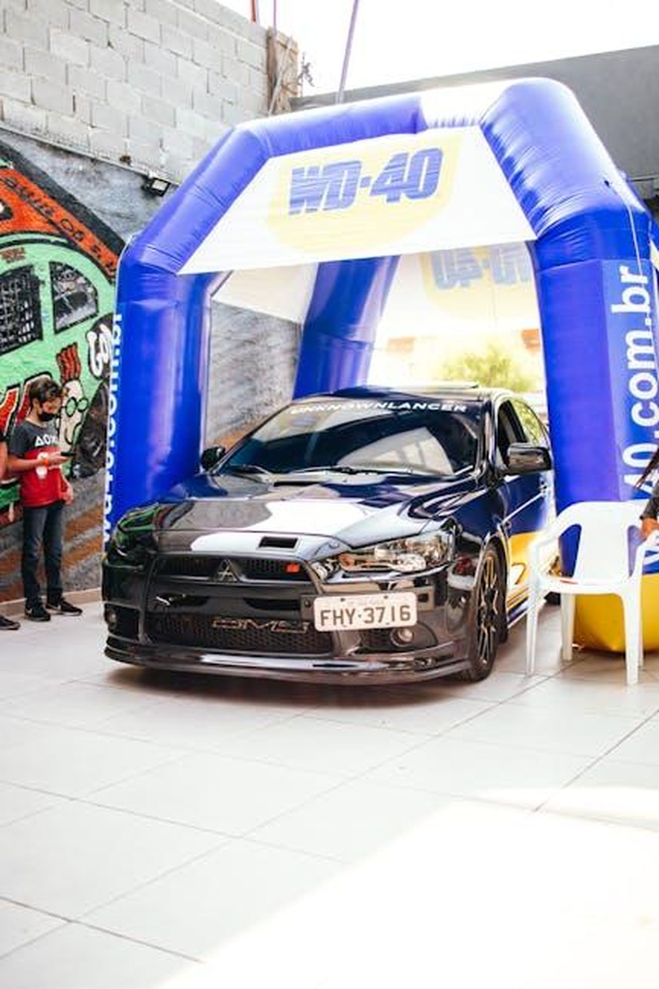 Promozioni_e_Offerte_Speciali_i7lREQbldF - Veicoli24 Source: Matheus Bertelli - pexels - Black race car showcased under a blue WD-40 inflatable arch at an outdoor event.