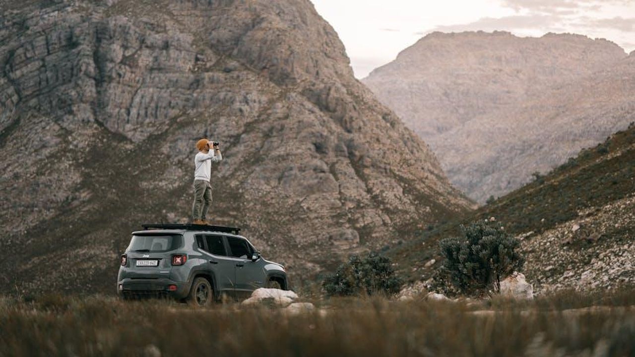 Renegade_e_Compass_edizione_speciale_North_Star_BEcAfUDVeT - Veicoli24 Source: Taryn Elliott - pexels - A man standing on an SUV roof using binoculars to view a mountain landscape.