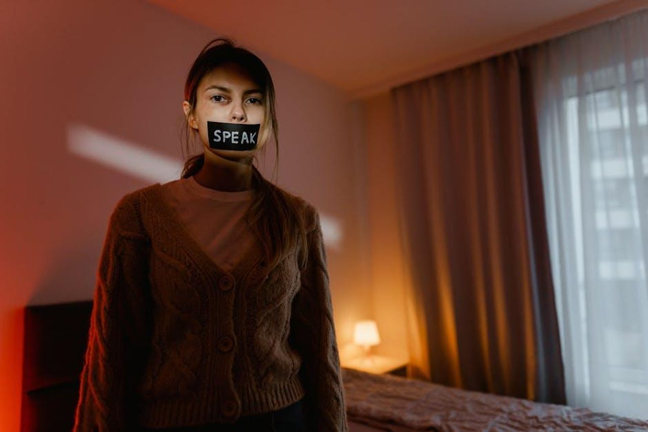 Un_evento_per_sensibilizzare_sulla_violenza_di_genere_9REJtLlGHL - Veicoli24 Source: MART PRODUCTION - pexels - A woman stands in a dimly lit bedroom with tape reading 'Speak' over her mouth, symbolizing silence and oppression.
